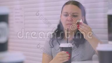 咖啡屋里的一个黑发女人从一个黑杯子里喝咖啡，吃一个蛋糕。 在玻璃上射击
